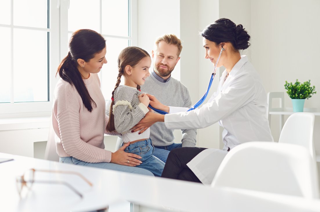 Pediatrician examining a smiling young girl's chest with a stethoscope while her parents sit beside her at Primary Med Clinic in Tucson, AZ